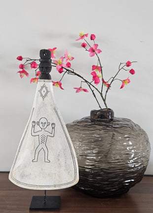 Zijaanzicht van ornament lepel wit naturel op zwarte standaard naast een donkere ronde vaas met roze bloesemtakken op een houten ondergrond
