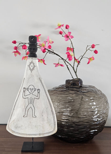 Zijaanzicht van ornament lepel wit naturel op zwarte standaard naast een donkere ronde vaas met roze bloesemtakken op een houten ondergrond