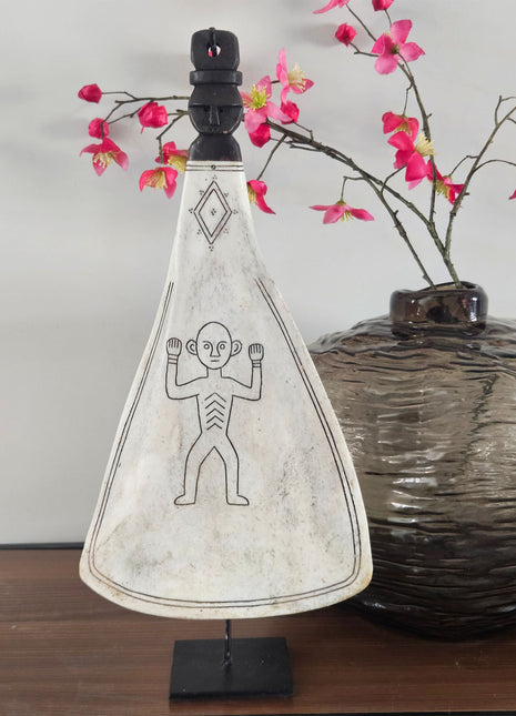 Ornament lepel wit naturel op zwarte standaard met gegraveerd figuur naast een donkere ronde vaas met roze bloesemtakken op een houten ondergrond