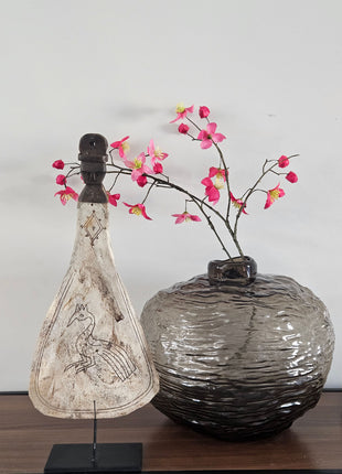 Lepel ornament naturel met gegraveerde pauw op een zwarte metalen standaard naast een grijze ronde vaas met roze bloesemtakken op een houten dressoir
