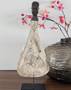 Lepel ornament naturel met gegraveerde pauw op een zwarte metalen standaard naast een grijze ronde vaas met roze bloesemtakken