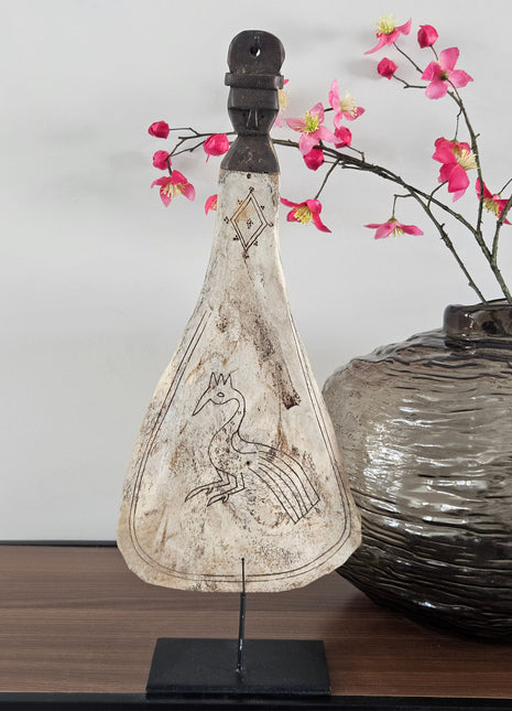Lepel ornament naturel met gegraveerde pauw op een zwarte metalen standaard naast een grijze ronde vaas met roze bloesemtakken