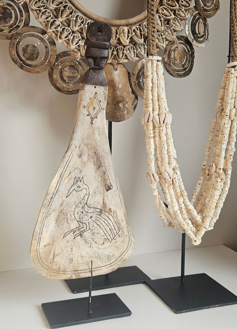 Ornament van een lepel op een standaard met daarin gegraveerd een pauw. Aan de rechtekant een witte kralenketting in ovale vorm met kleine witte schelpen.