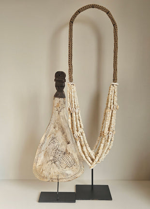 Ornament van een lepel op een standaard met daarin gegraveerd een pauw. Aan de rechtekant een witte kralenketting in ovale vorm met kleine witte schelpen.