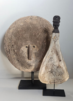 Een ornament in de vorm van een lepel met daarin gegraveerd de afbeelding van een pauw. Daarachter een gezicht uitgesneden uit hout in de vorm van een masker.