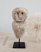 Close-up van stenen poppetje beige met gegraveerd gezicht op een zwarte metalen standaard op een linnen ondergrond naast een witte gedroogde paardenbloem