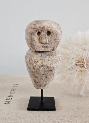 Close-up van stenen poppetje beige met gegraveerd gezicht op een zwarte metalen standaard op een linnen ondergrond naast een witte gedroogde paardenbloem