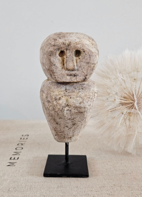 Close-up van stenen poppetje beige met gegraveerd gezicht op een zwarte metalen standaard op een linnen ondergrond naast een witte gedroogde paardenbloem