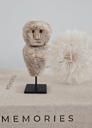 Stenen poppetje beige op zwarte metalen standaard op een linnen ondergrond naast een witte gedroogde paardenbloem