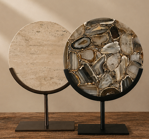 Twee ronde schijfornamenten op zwarte metalen standaarden op een houten ondergrond: links een beige travertijn schijf, rechts een grotere schijf met grijze en bruine agaatplakken met gouden rand

