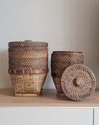 Grote en kleine rieten opbergmand bruin met deksel naast elkaar op een houten dressoir waarvan de kleine mand op zijn kant ligt met deksel ernaast