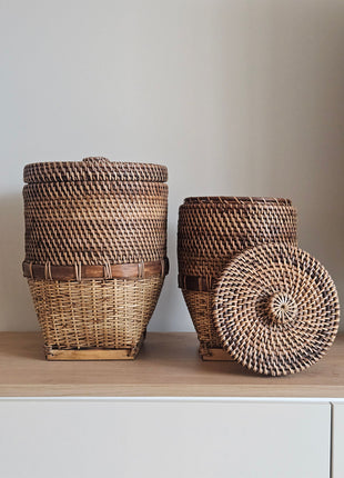 Grote en kleine rieten opbergmand bruin met deksel naast elkaar op een houten dressoir waarvan de kleine mand op zijn kant ligt met deksel ernaast