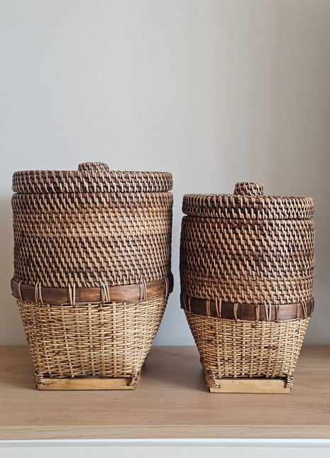 Grote en kleine rieten opbergmand bruin met deksel naast elkaar op een houten dressoir op een lichte achtergrond