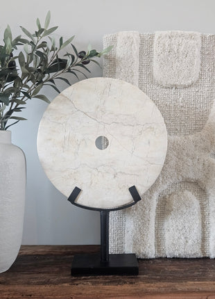 Rond marmeren ornament wit op een zwarte metalen standaard naast een witte vaas met olijftakken op een houten ondergrond