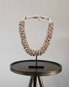 Schelpenketting beige M op zwarte metalen standaard met beige kauri-schelpen en gevlochten witte band op een donkere ronde bijzettafel voor een witte muur