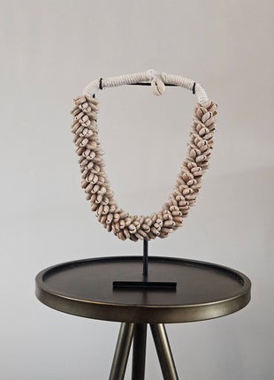 Schelpenketting beige M op zwarte metalen standaard met beige kauri-schelpen en gevlochten witte band op een donkere ronde bijzettafel voor een witte muur