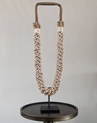 Schelpenketting beige L op zwarte metalen standaard met beige kauri-schelpen en gevlochten witte band op een witte achtergrond