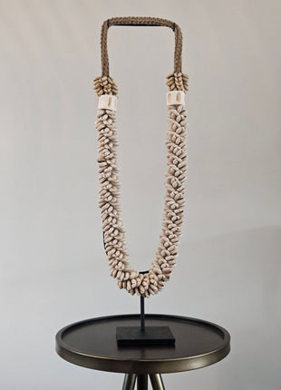 Schelpenketting beige L op zwarte metalen standaard met beige kauri-schelpen en gevlochten witte band op een witte achtergrond