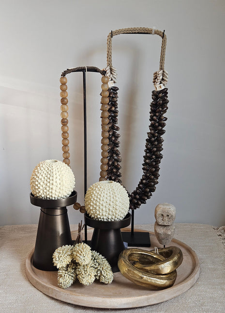 Schelpenketting bruin L op zwarte standaard naast een lichtbruine kralenketting op standaard twee witte schelpenbolornamentjes en een gouden ring-ornament op een houten rond dienblad op een linnen ondergrond