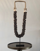 Schelpenketting bruin L op zwarte metalen standaard met donkerbruine ovale schelpen en gevlochten jute band op een witte achtergrond