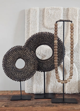 Schelpenketting bruin L op zwarte standaard naast een bronzen kralenketting op standaard op een houten dressoir voor een witte muur