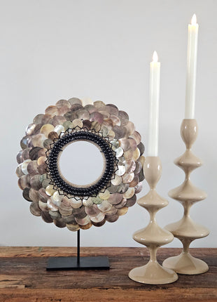 Schelpenketting gepolijste schelpen L op zwarte standaard naast twee taupe keramieke kandelaars op een houten dressoir voor een crème schilderij met boogmotief