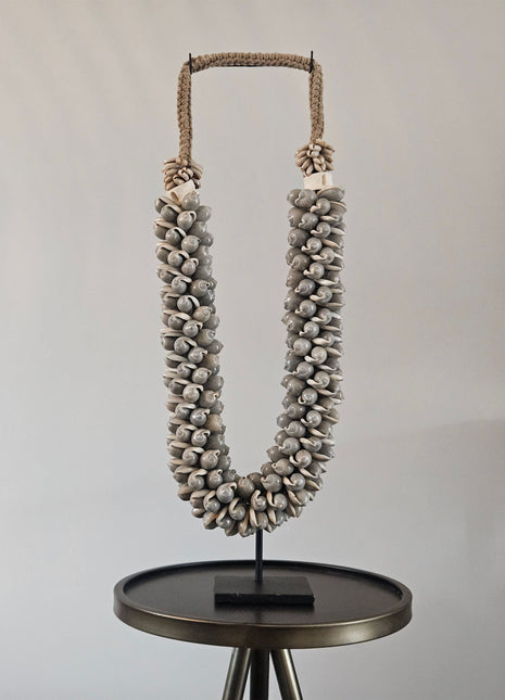 Schelpenketting grijs op zwarte metalen standaard met ronde grijze schelpen en witte kauri-schelpen op een gevlochten jute band op een donkere ronde bijzettafel