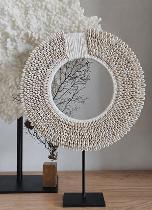 Schelpenketting wit-beige L op zwarte metalen standaard op een houten dressoir naast een zwarte houten kam op standaard en een beige onyx schijfornament op standaard