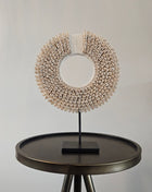 Schelpenketting wit S op zwarte metalen standaard met kauri-schelpen en gevlochten witte band op een witte achtergrond
