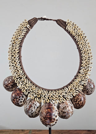 Schelpenketting tijgerschelp L op zwarte metalen standaard met kauri-schelpen gevlochten jute band en grote tijgerschelpen als hanger op een lichtgrijze achtergrond