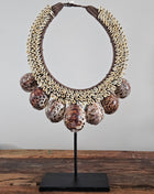 Schelpenketting tijgerschelp L op zwarte metalen standaard met kauri-schelpen gevlochten jute band en grote tijgerschelpen als hanger op een houten dressoir naast een wit koraalornament en een bronzen vaas