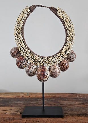 Schelpenketting tijgerschelp L op zwarte metalen standaard met kauri-schelpen gevlochten jute band en grote tijgerschelpen als hanger op een houten dressoir naast een wit koraalornament en een bronzen vaas