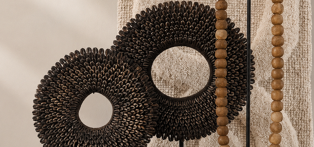 Close-up van twee donkerbruine schelpenkettingen op zwarte standaarden voor een geweven wandpaneel in naturel, met een houten kralenketting op een standaard op de achtergrond

