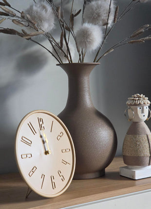 Schuin vooraanzicht van beige Liva tafelklok met gladde afwerking naast een taupe bloemenvaas en Namji pop op hout