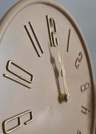 Schuine close-up van beige Liva tafelklok met gladde afwerking en goudkleurige wijzers en cijfers tegen grijs