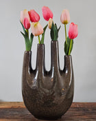 Tulpenvaas bruin speckled aardewerk met vier hoge halsopeningen en roze-rode tulpen op een houten ondergrond voor een lichtgrijze muur