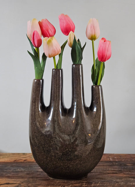 Tulpenvaas bruin speckled aardewerk met vier hoge halsopeningen en roze-rode tulpen op een houten ondergrond voor een lichtgrijze muur