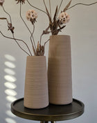 Vooraanzicht van twee beige Elora vazen met ruwe afwerking en creme kunstbloemen op een rond metalen tafeltje