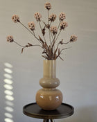 Vooraanzicht van taupe Livana vaas met glanzende afwerking en creme kunstbloemen op een rond zwart tafeltje