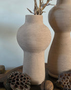 Vooraanzicht van taupe Marconne vaas met ruwe afwerking naast een grotere vaas en lotus op een houten dienblad