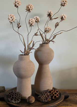 Vooraanzicht van twee taupe Marconne vazen met ruwe afwerking en beige kunstbloemen naast lotus op houten dienblad