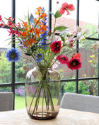 Vrijstaande vaas Artic M van Vase the World in glanzend taupe glas gestileerd met kleurijke bloemen op een tafel met op de achtergond een kleurrijke tuin.