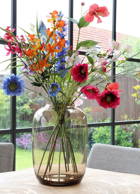 Vrijstaande vaas Artic M van Vase the World in glanzend taupe glas gestileerd met kleurijke bloemen op een tafel met op de achtergond een kleurrijke tuin.
