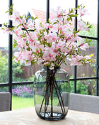 Vrijstaande vaas Artic M van Vase the World in glanzend taupe glas gestileerd met kleurijke bloemen op een tafel met op de achtergond een kleurrijke tuin.