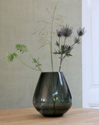 Vrijstaande druppelvormige vaas Tasman M van Vase the World in glanzend taupe glas gestileerd met kleurijke bloemen op een tafel.