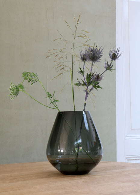 Vrijstaande druppelvormige vaas Tasman M van Vase the World in glanzend taupe glas gestileerd met kleurijke bloemen op een tafel.