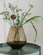 Vrijstaande druppelvormige vaas Tasman M van Vase the World in glanzend taupe glas gestileerd met kleurijke bloemen op een sidetable.