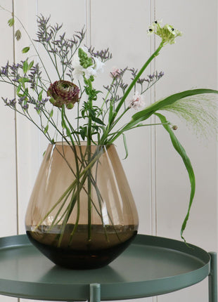 Vrijstaande druppelvormige vaas Tasman M van Vase the World in glanzend taupe glas gestileerd met kleurijke bloemen op een sidetable.
