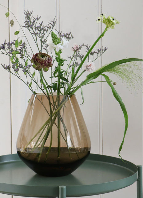 Vrijstaande druppelvormige vaas Tasman M van Vase the World in glanzend taupe glas gestileerd met kleurijke bloemen op een sidetable.
