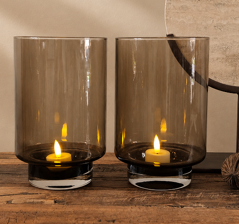 Twee rookglazen windlichten met brandende waxinelichten op een houten ondergrond, met op de achtergrond een rond ornament en een droogtak

